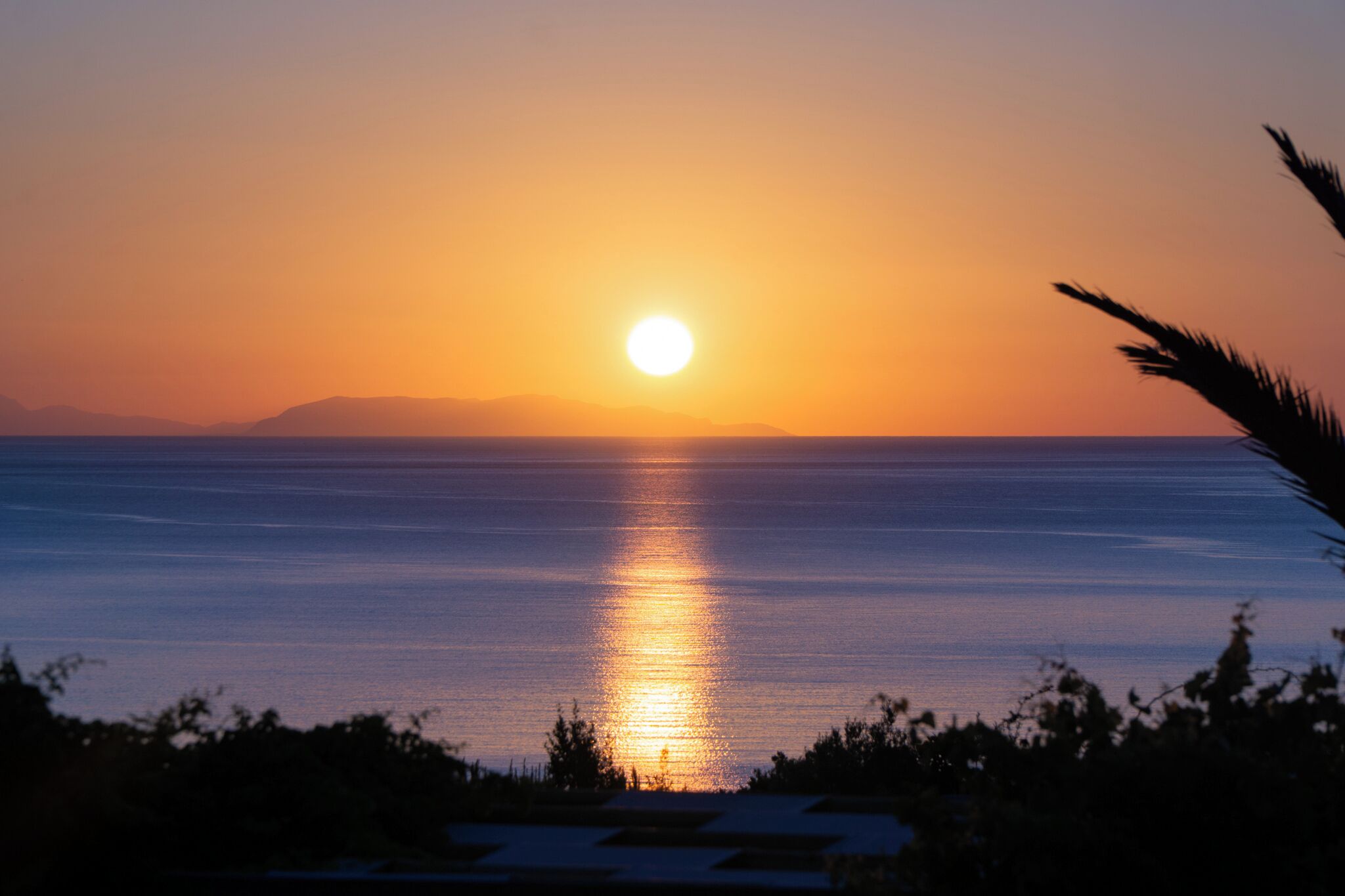 A sunset over a calm sea with silhouettes of trees and a distant mountain range.
