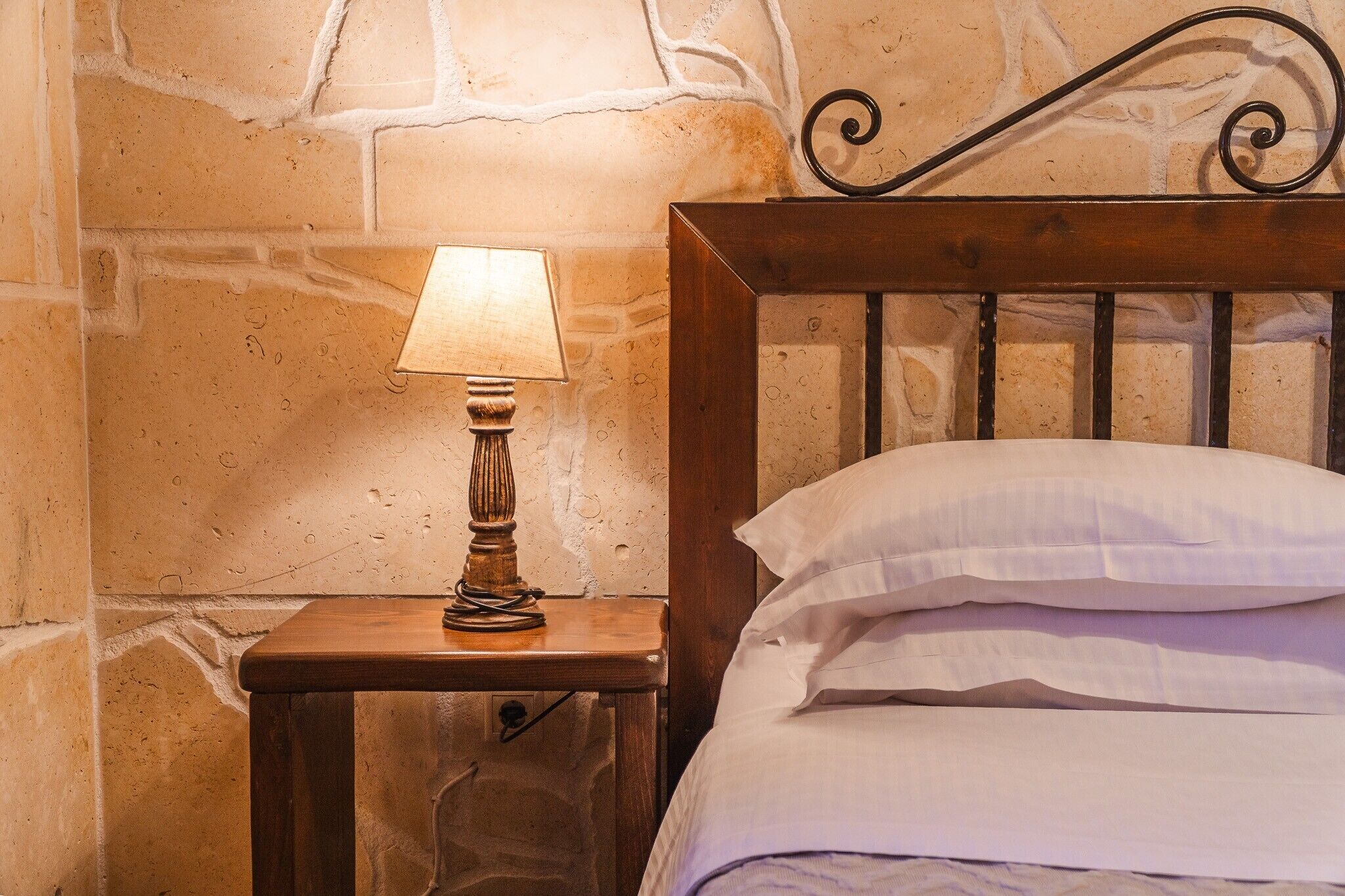 A wooden bedside table with a lamp and a bed with white linens.