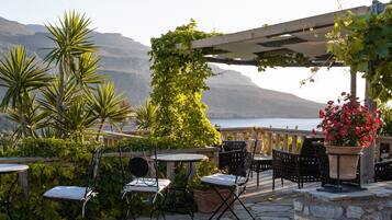 An outdoor patio with tables and chairs, surrounded by greenery and a view of a body of water and mountains.