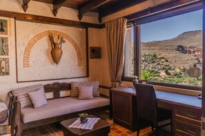 A room with a sofa, a desk, and a view of a desert landscape. - Terra Minoika Resort (Sitia)
