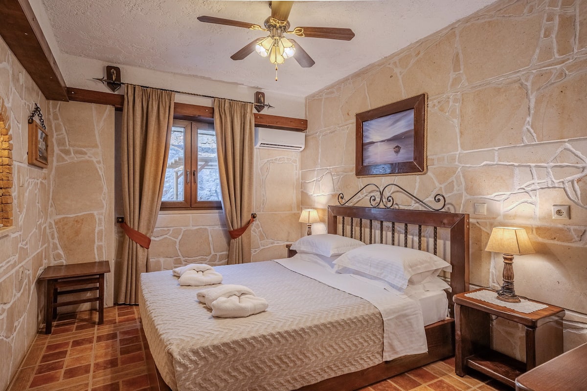 A bedroom with a stone wall, a wooden bed frame, a ceiling fan, and a window with curtains.
