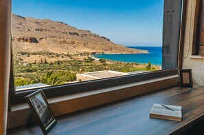 A room with a view of a coastal landscape, a wooden desk with a book and a picture frame, and a window with a wooden frame. - Terra Minoika Resort (Sitia)