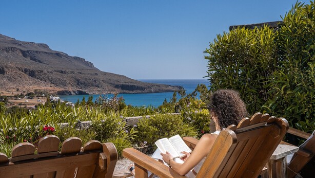 Una persona leyendo un libro en una silla de madera con vista a un paisaje costero.