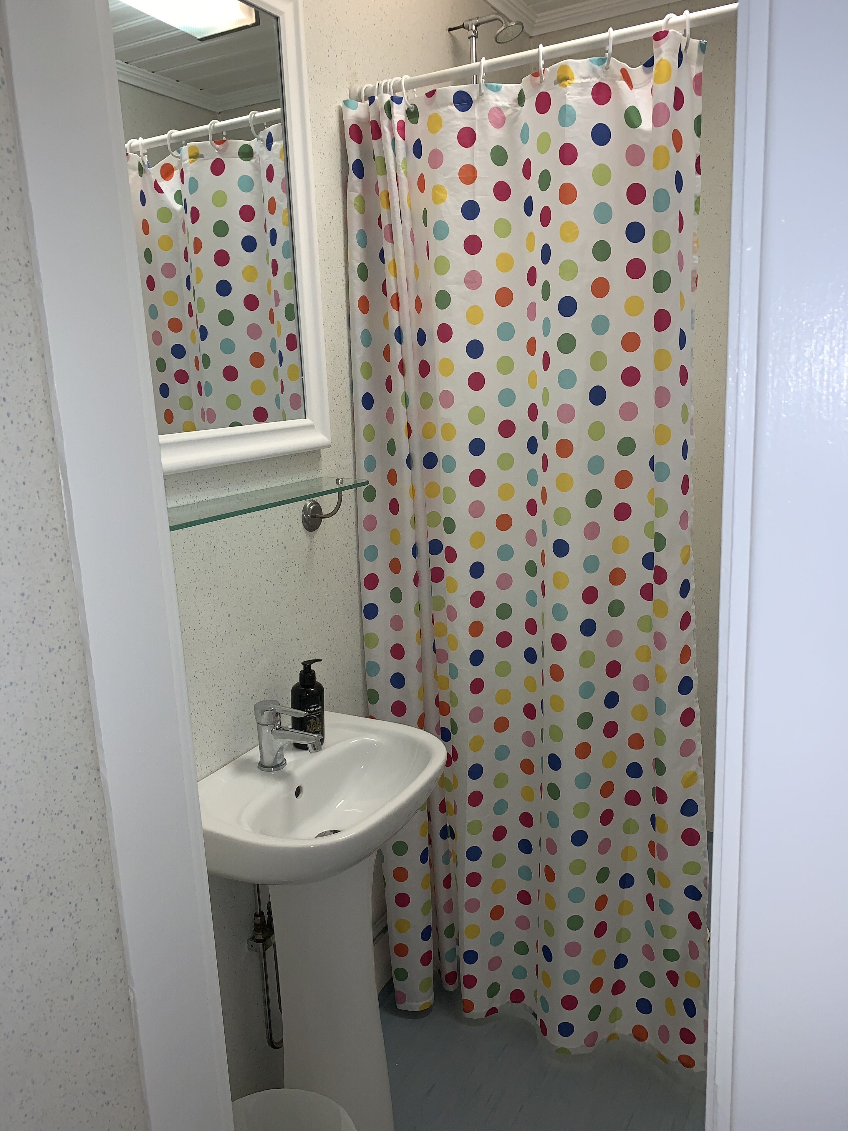 Une salle de bain avec un lavabo blanc, une étagère en verre au-dessus et un rideau de douche à pois colorés.