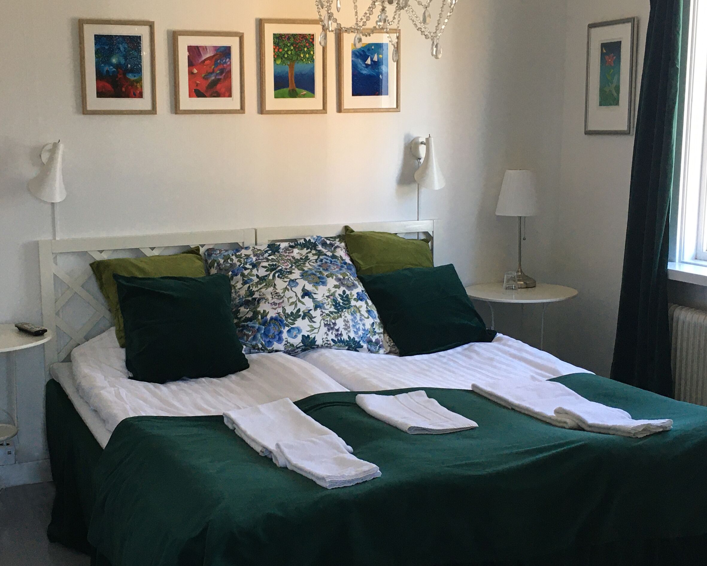 Una cama bien hecha con ropa de cama verde, dos mesitas de noche con lámparas y cuatro cuadros enmarcados en la pared.