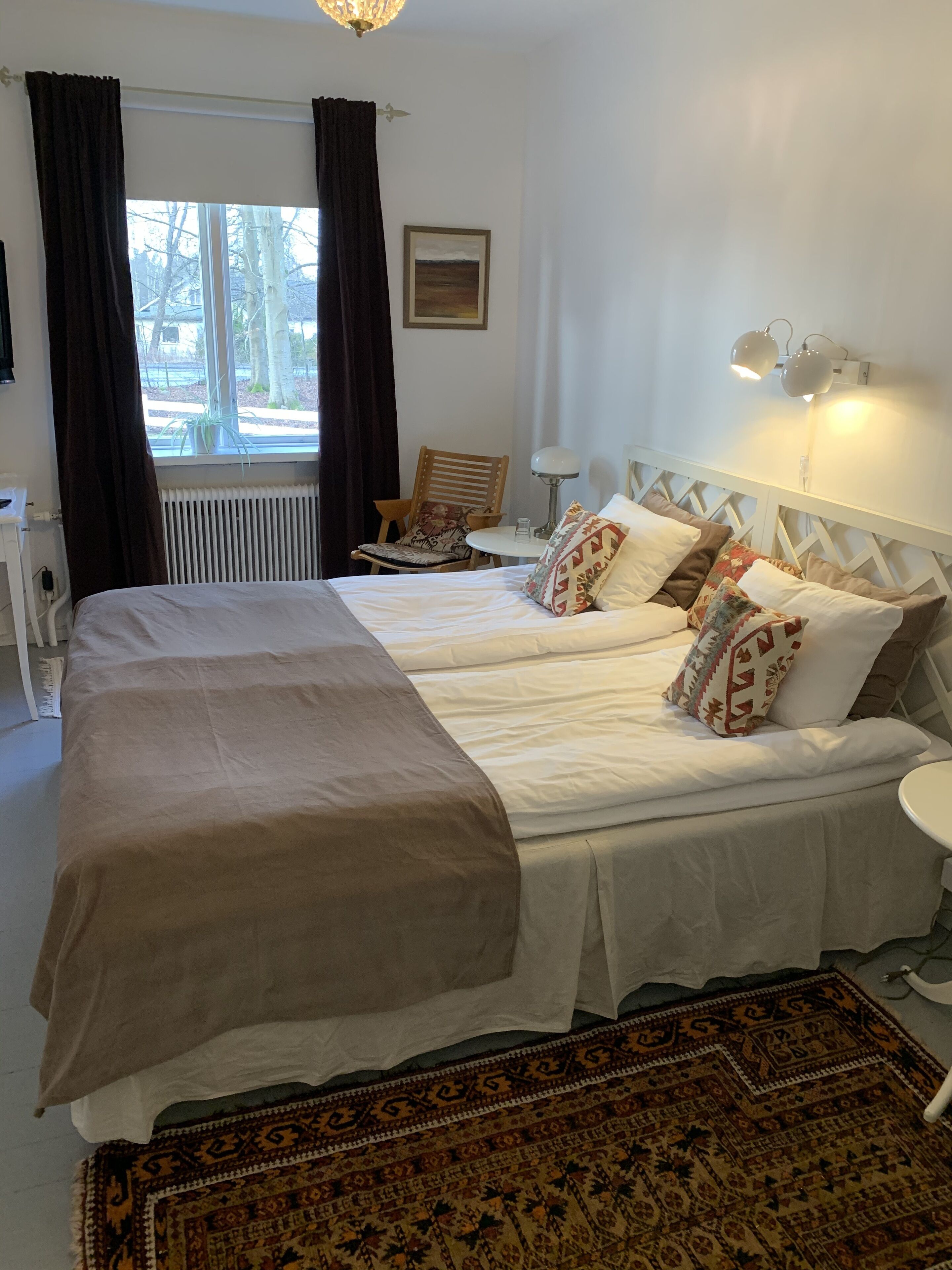 Una cama doble con cubrecama color beige, una alfombra estampada, una silla con cojín floral y una mesita con lámpara.