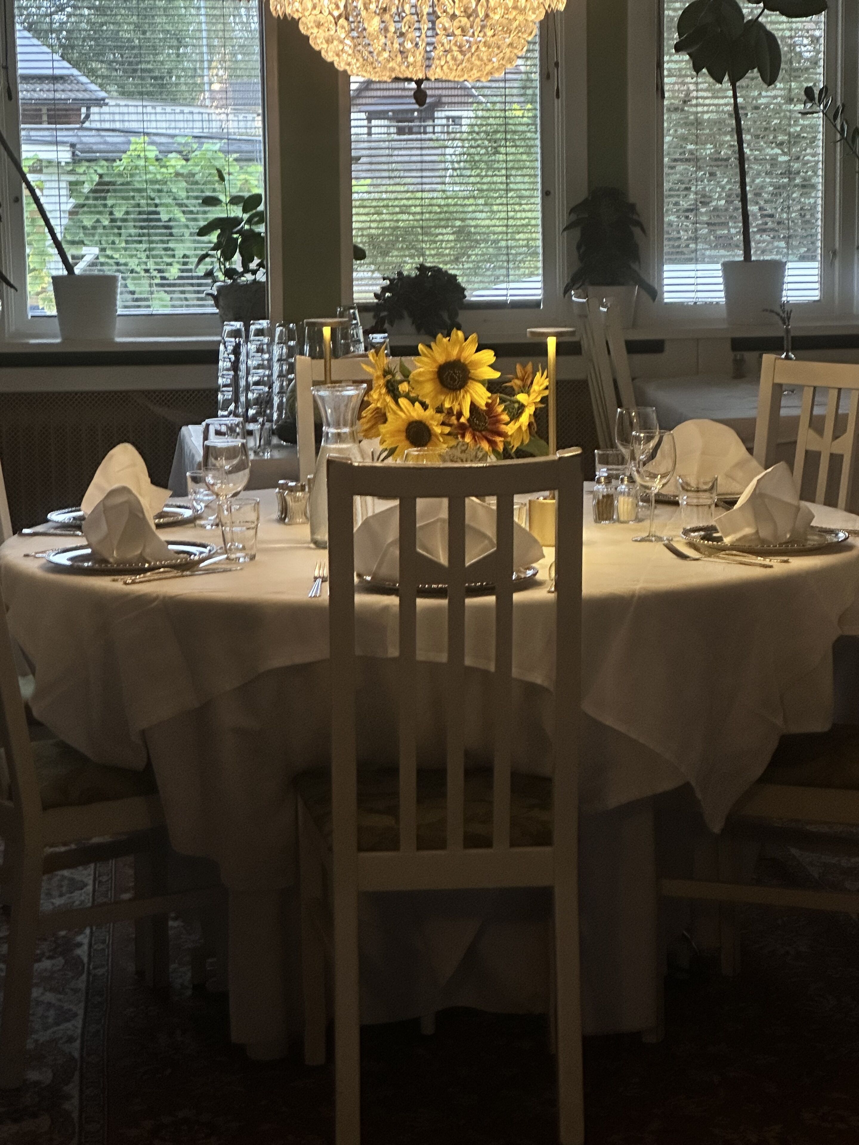 A dining table set for four with white tablecloths, sunflowers, and glassware.
