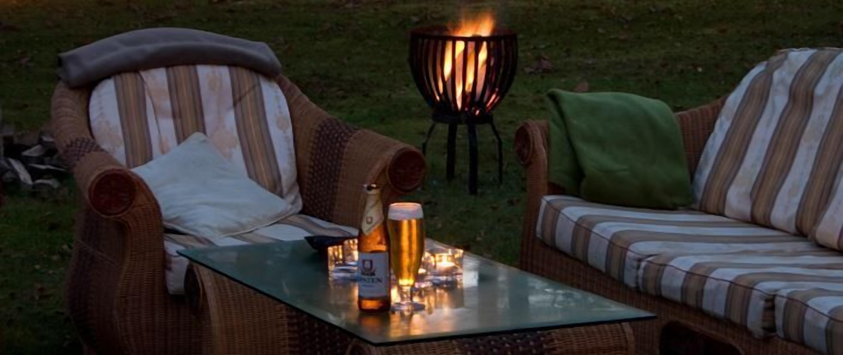 A cozy outdoor seating area with wicker chairs and a glass table, a fire pit in the background, and a bottle of beer and a glass on the table.