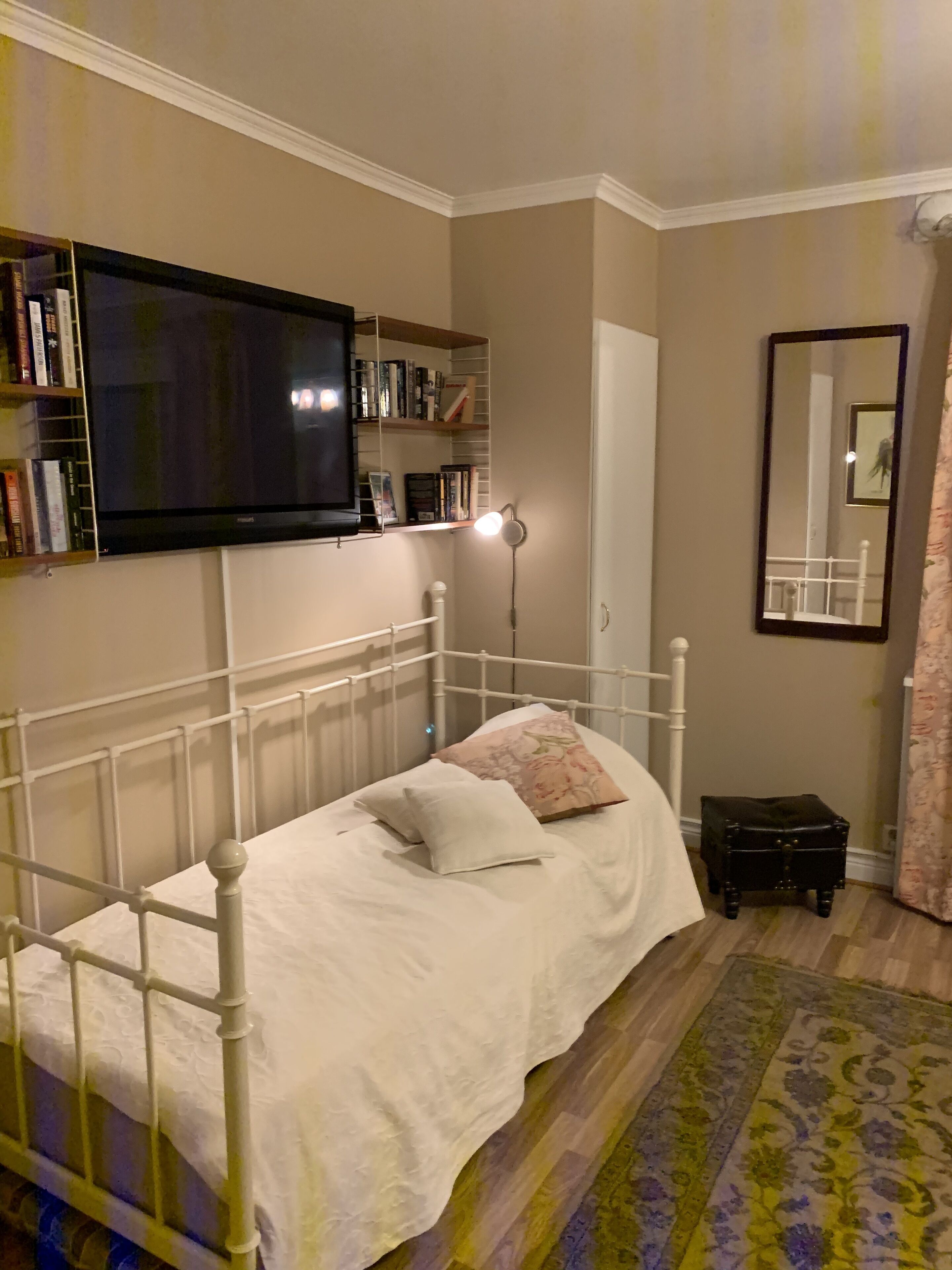 Une chambre à coucher avec un lit en métal blanc, une table de chevet, une petite table de nuit noire, un téléviseur fixé au mur et une bibliothèque contenant des livres et des objets décoratifs.