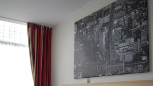 Une chambre d’hôtel avec un grand lit, une tête de lit en bois, une table de chevet, une chaise, une fenêtre avec des rideaux et un tableau représentant un paysage urbain accroché au mur.