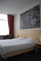 Quarto de hotel com uma cama grande, cabeceira de madeira, mesa de cabeceira, uma cadeira, janela com cortinas e uma pintura de paisagem urbana na parede.