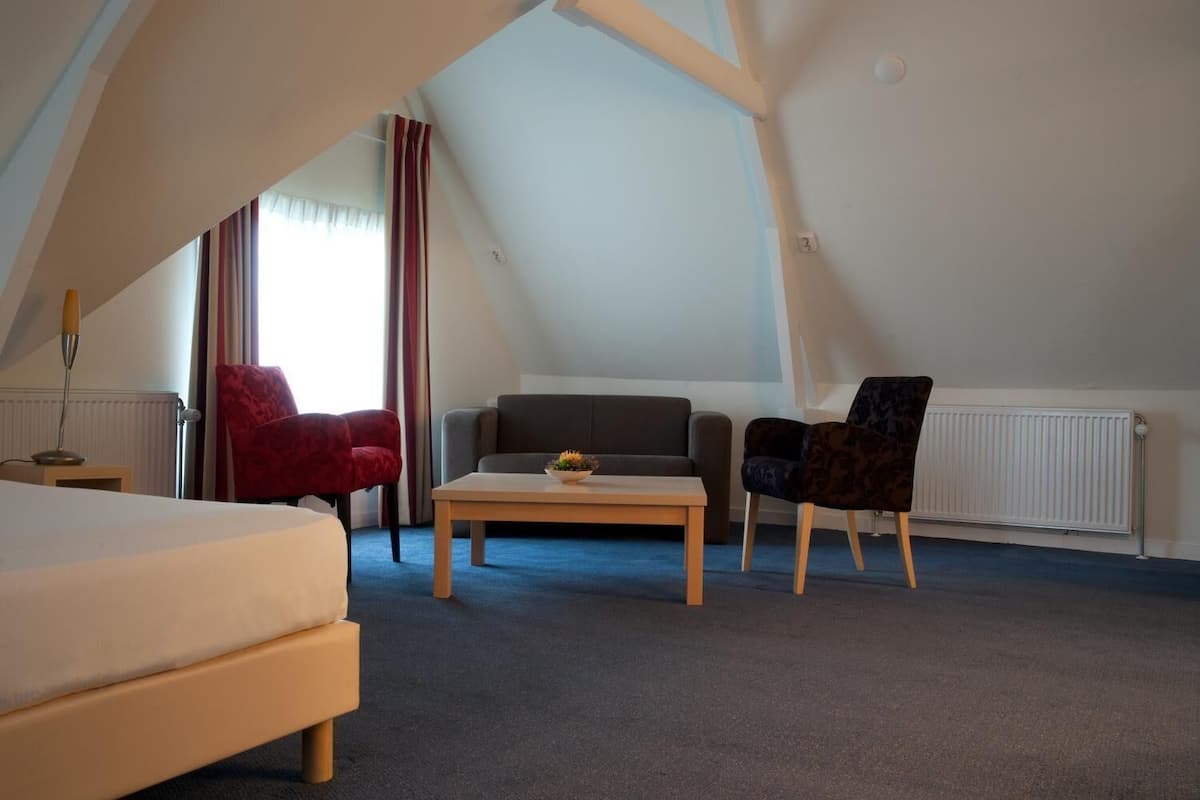 Una habitación con una cama, un sillón rojo, un sofá gris, un sillón estampado oscuro y una mesa de centro de madera.