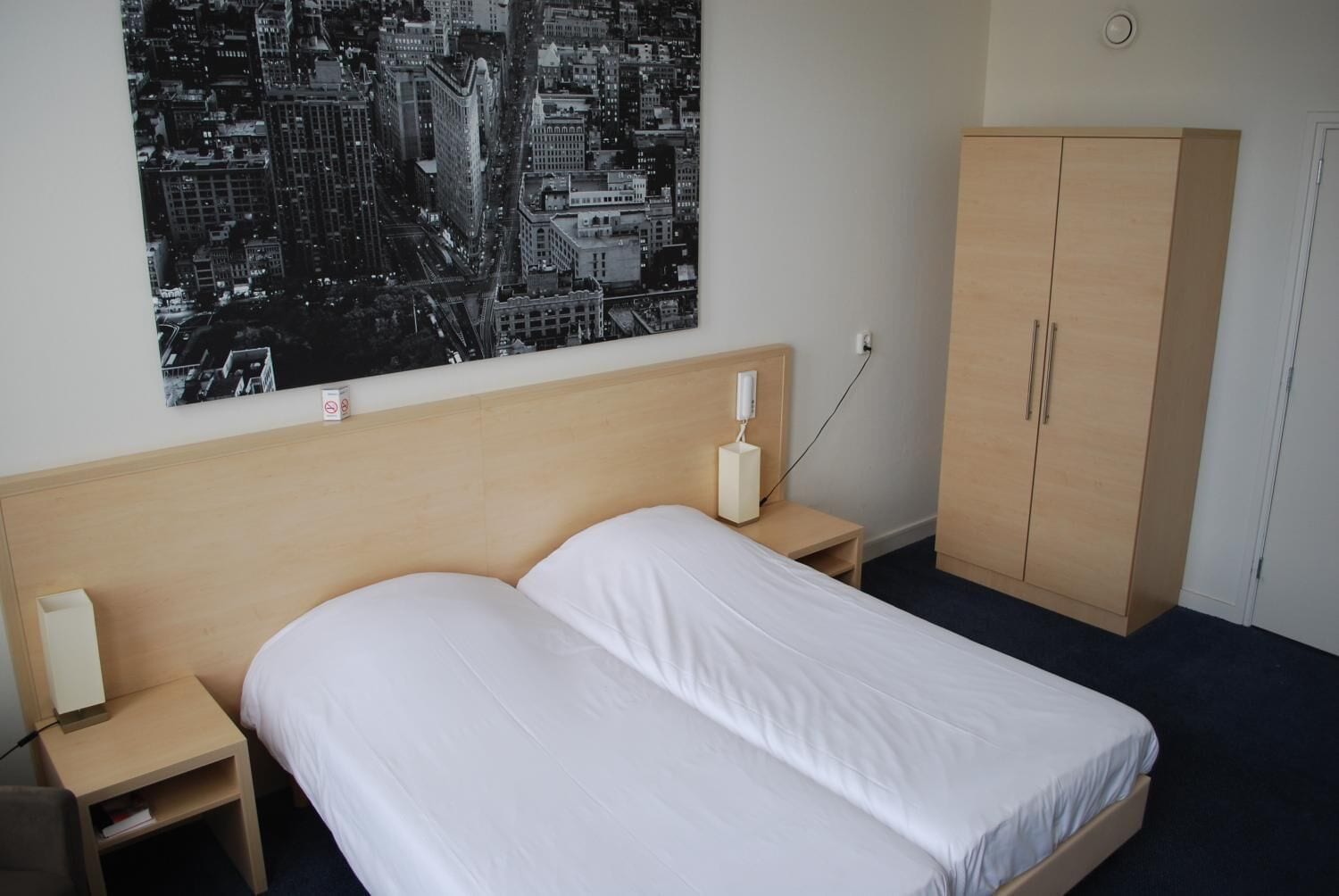 A hotel room with a bed, a nightstand, a bedside lamp, a wardrobe, and a cityscape wall art.