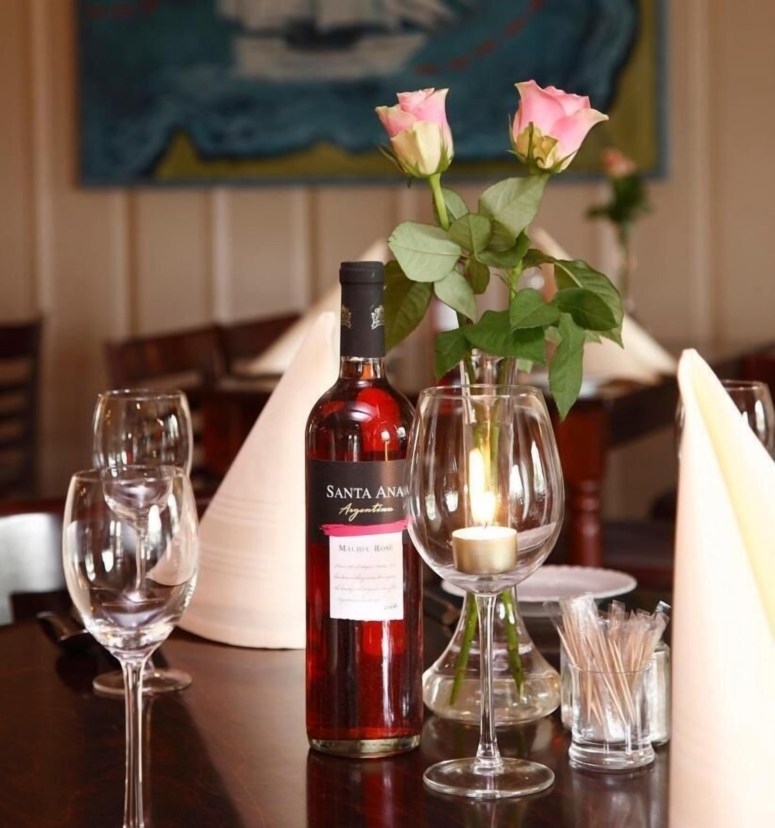 Una mesa con una botella de vino Santa Ana, dos copas de vino vacías, una vela y un florero con rosas.