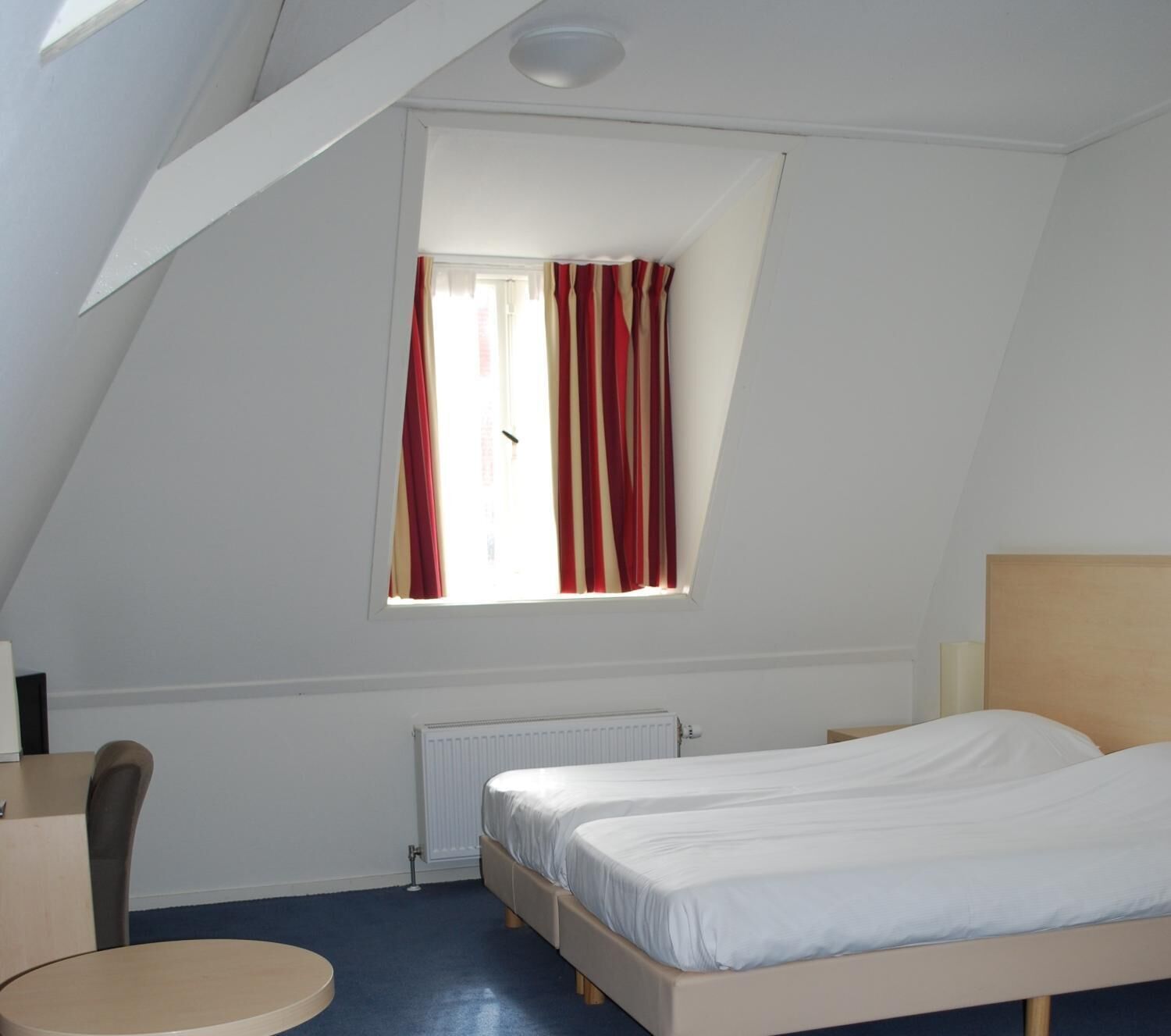 A bedroom with a sloped ceiling, a window with red curtains, a bed, a chair, and a radiator.