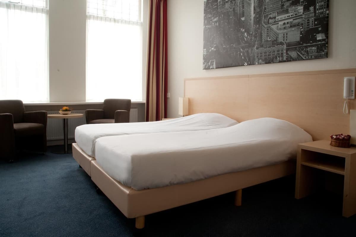 Una habitación de hotel con una cama, dos sillas, una mesita con un florero, una lámpara y un cuadro de un paisaje urbano.