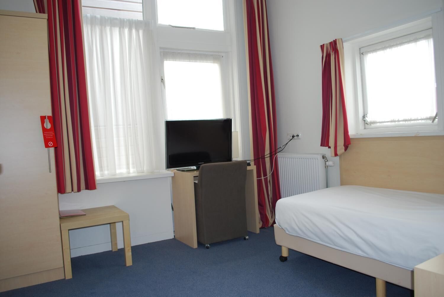 A hotel room with a bed, a desk with a computer, a chair, a small table, and a radiator.