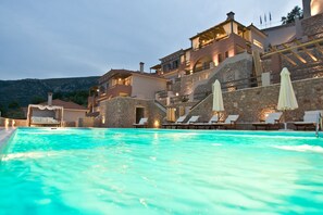 A pool area with lounge chairs and umbrellas, surrounded by stone buildings with multiple balconies and a mountainous backdrop. - Apelon Tiritas Villas (South Kynouria)