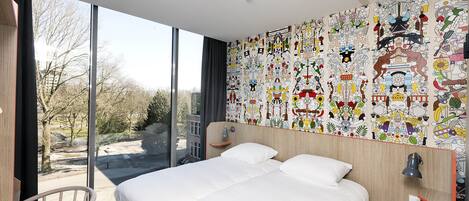 A modern hotel room with a large bed, a chair, and a view of the outdoors through floor-to-ceiling windows.