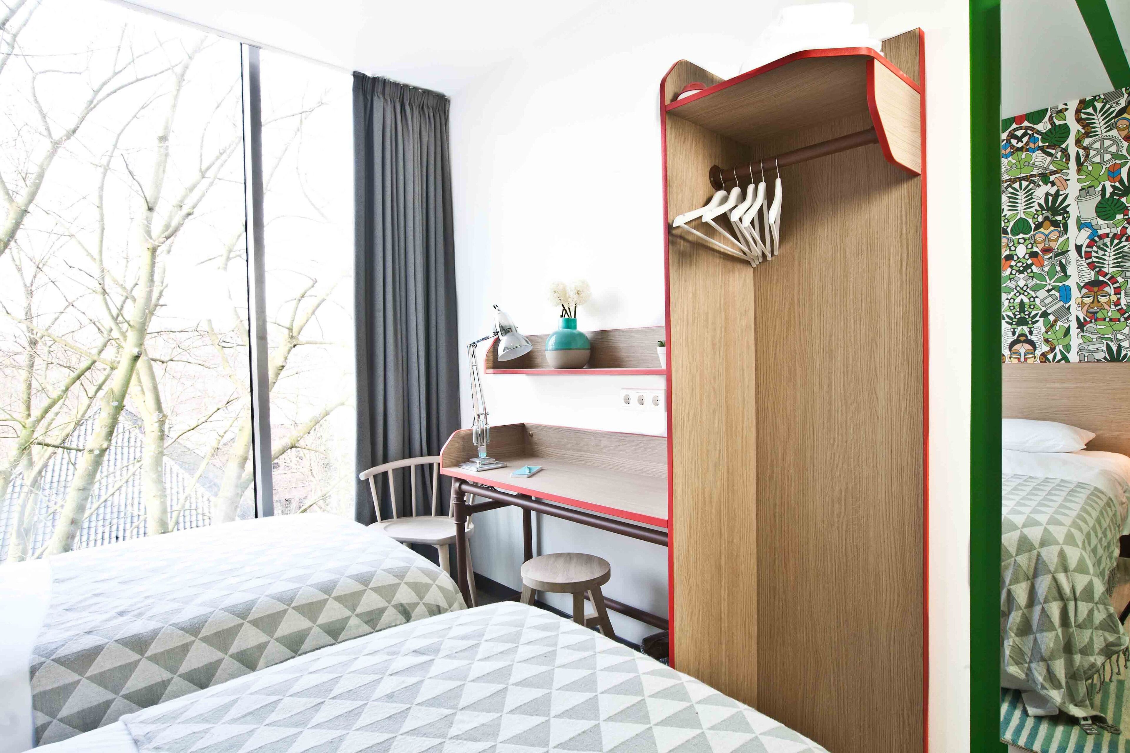 Ein Hotelzimmer mit zwei Betten, einem Schreibtisch und einem Stuhl. Ein Fenster mit Blick auf Bäume und einem Vorhang.