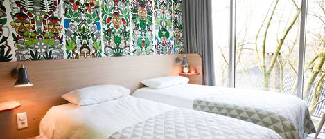 A hotel room with two beds, a wooden headboard, a nightstand with a lamp, and a wall with a colorful mural.