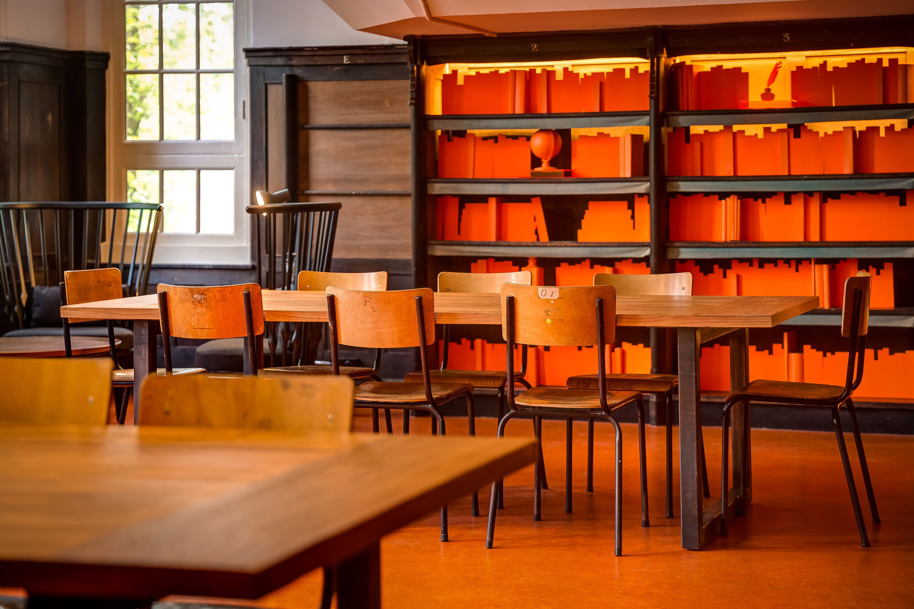 Eine Bibliothek mit Holztischen und -stühlen, einem großen Fenster und orangefarbenen Regalen.