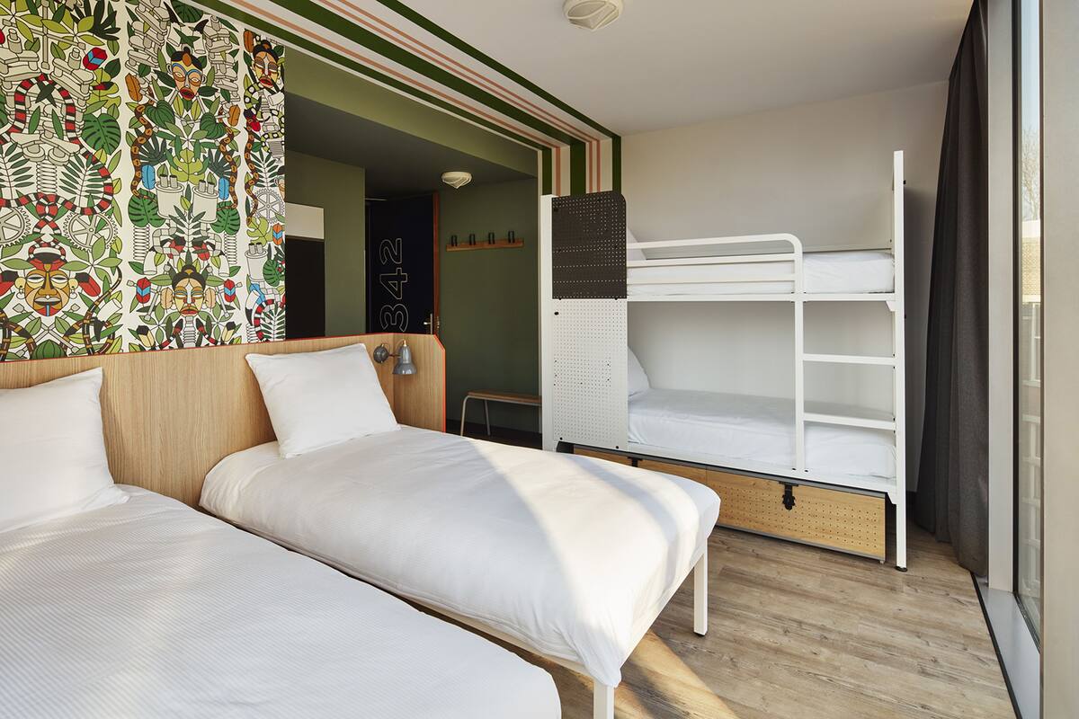 Une chambre d’hôtel avec deux lits simples, un lit superposé, une tête de lit en bois, un mur à motifs et une fenêtre avec des rideaux.