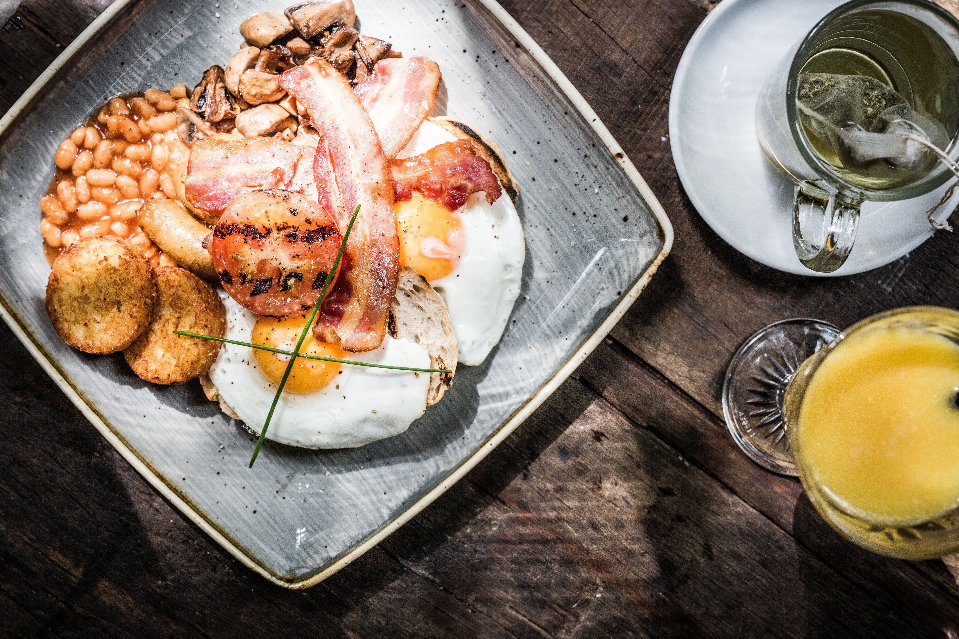 Ein Frühstücksteller mit Eiern, Speck, Bohnen, Pilzen und Toast auf einer Keramikplatte.