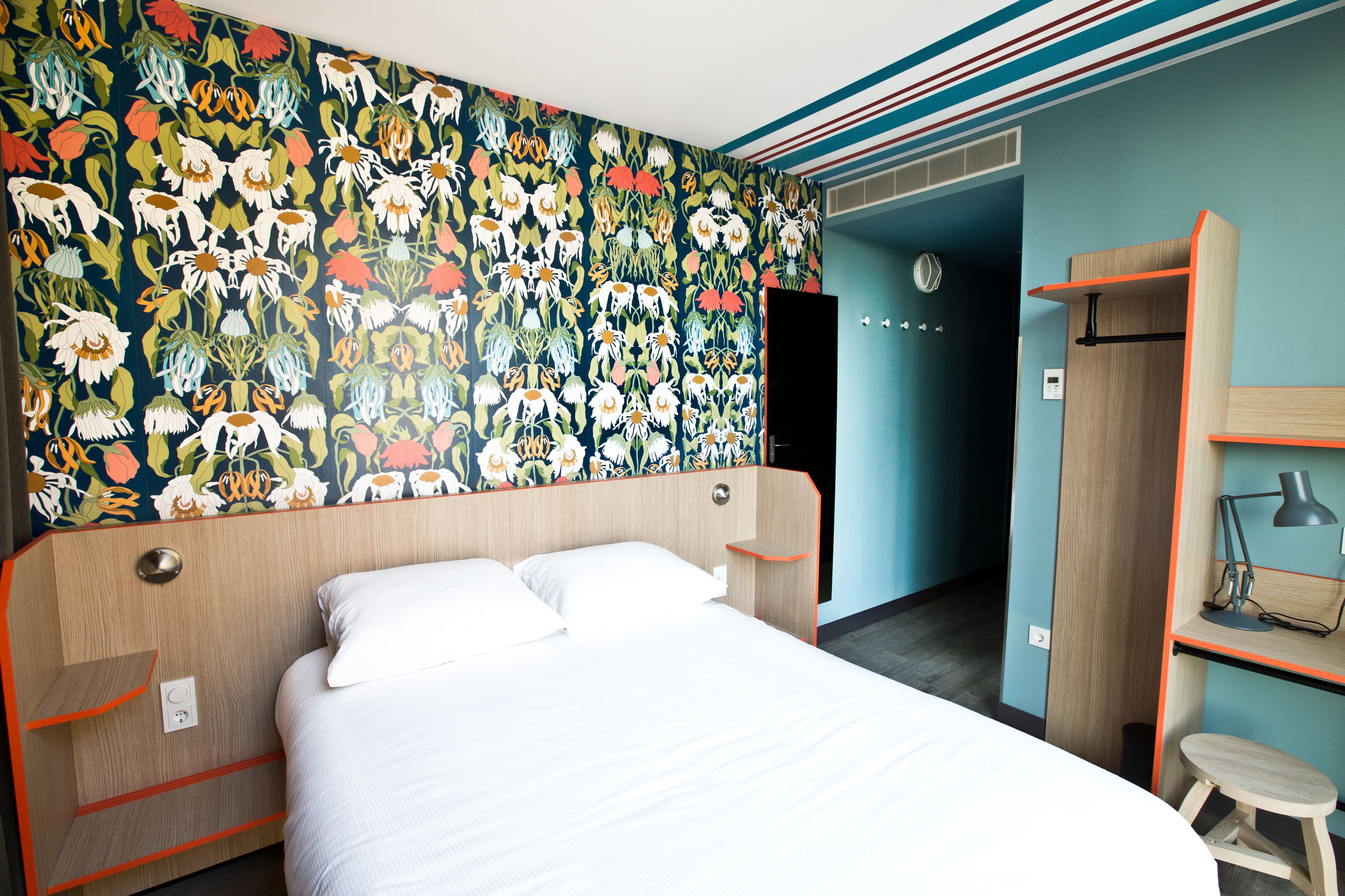 A hotel room with a bed, a wardrobe, a desk, and a colorful floral wallpaper.