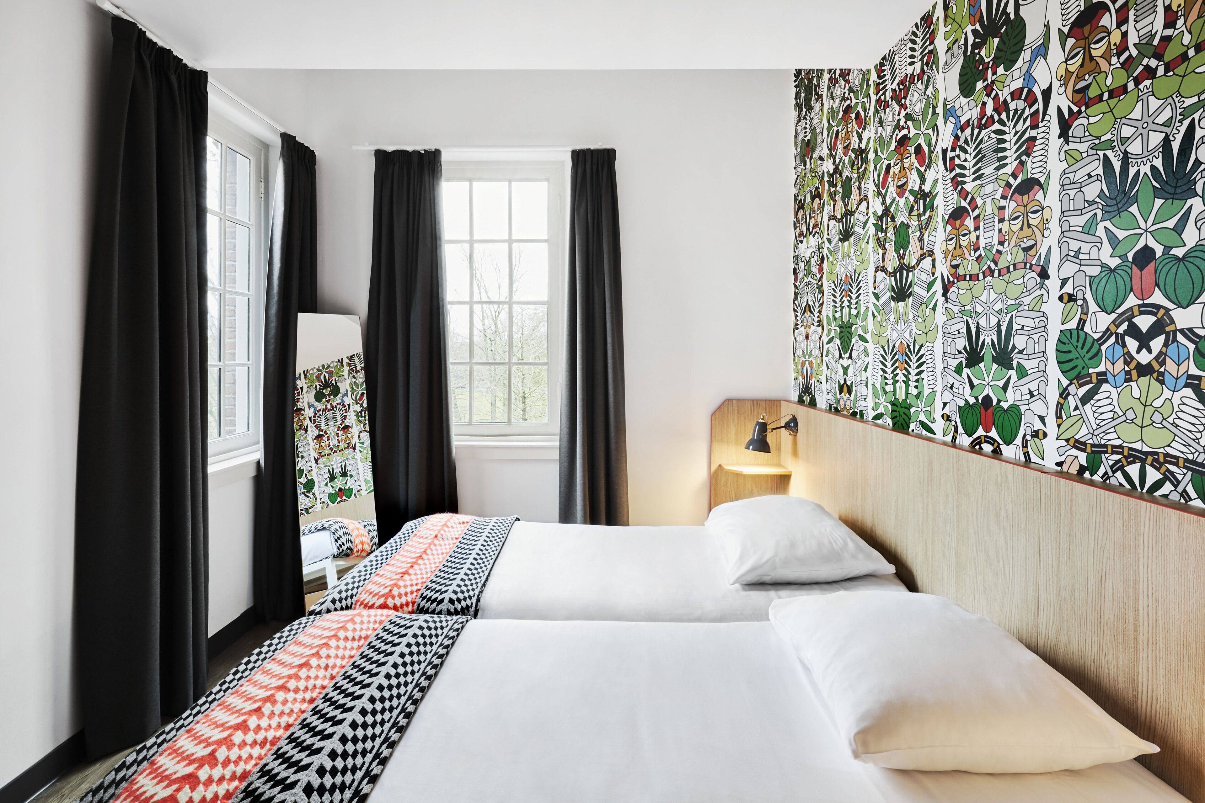 A hotel room with two beds, black curtains, a window, a wall with a colorful pattern, and a bedside table with a lamp.