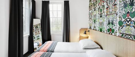 A hotel room with two beds, black curtains, a window, a wall with a colorful pattern, and a bedside table with a lamp.
