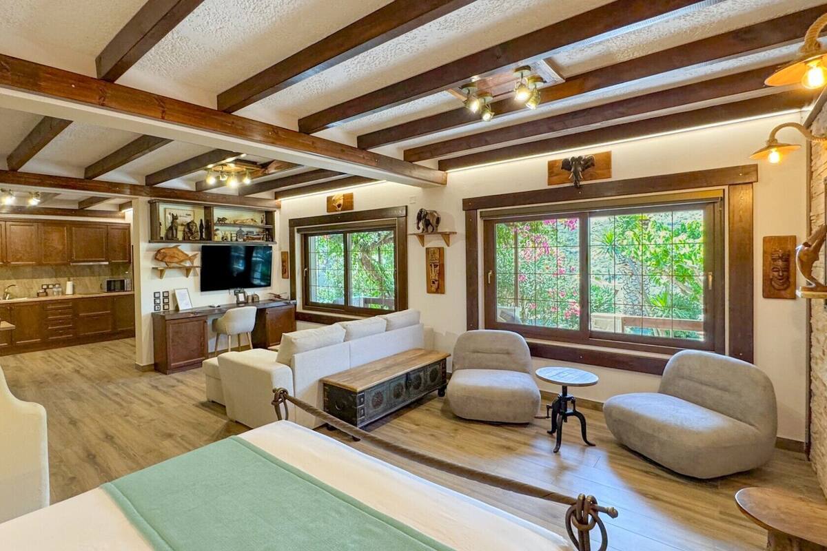 A spacious living area with a large window, wooden beams, a television, and a kitchen in the background.