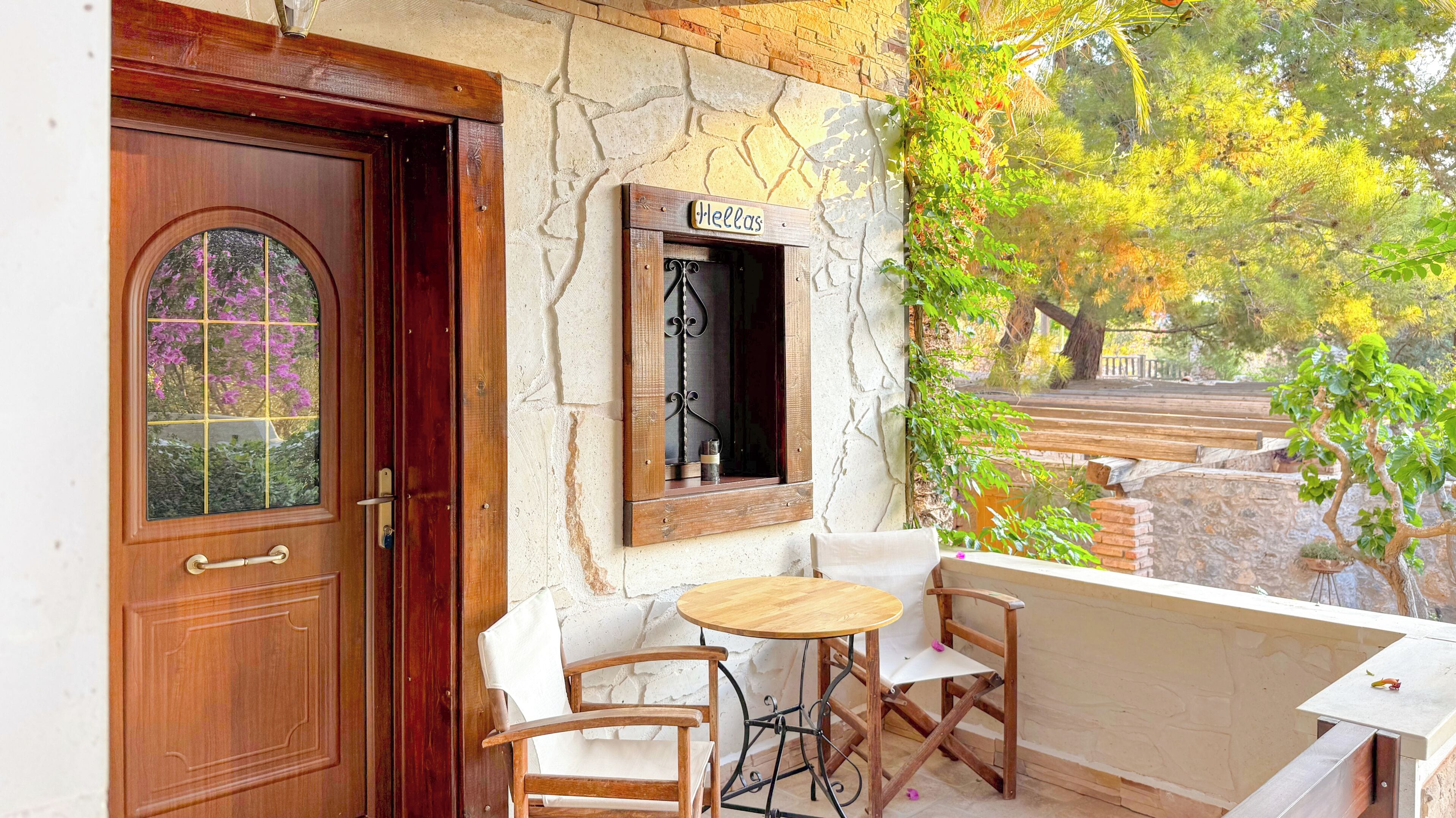 Una puerta de madera con un panel de vidrio, una pequeña ventana con un cartel y una mesa con dos sillas frente a un edificio de paredes de piedra.