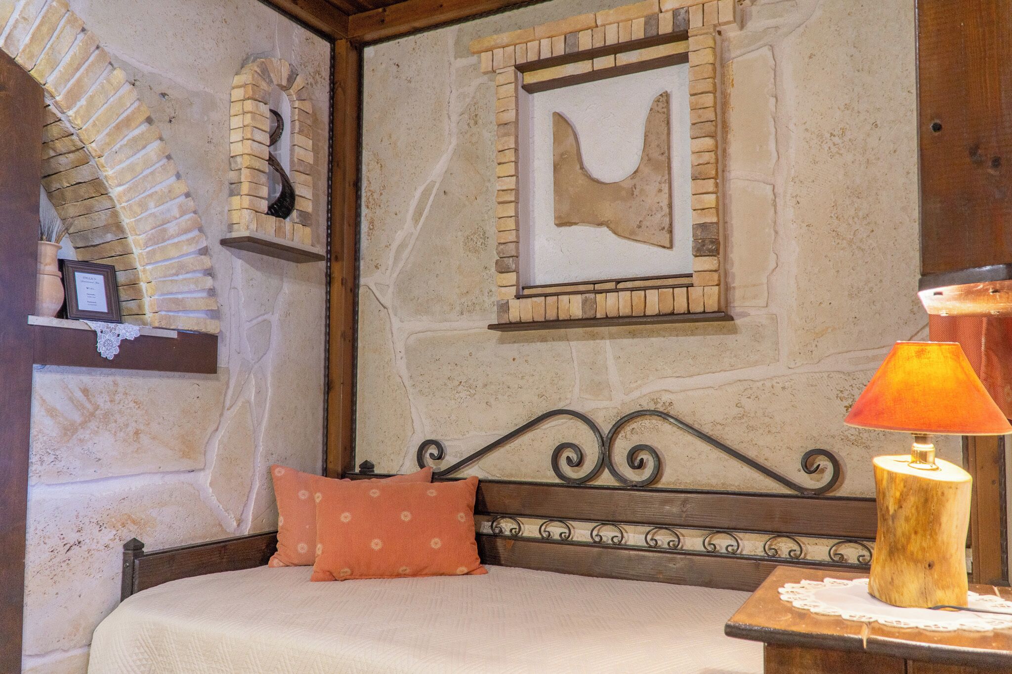 Una habitación con pared de piedra, una cama con cabecera, una mesita de noche de madera, un cuadro colgado en la pared y un pequeño estante con adornos.