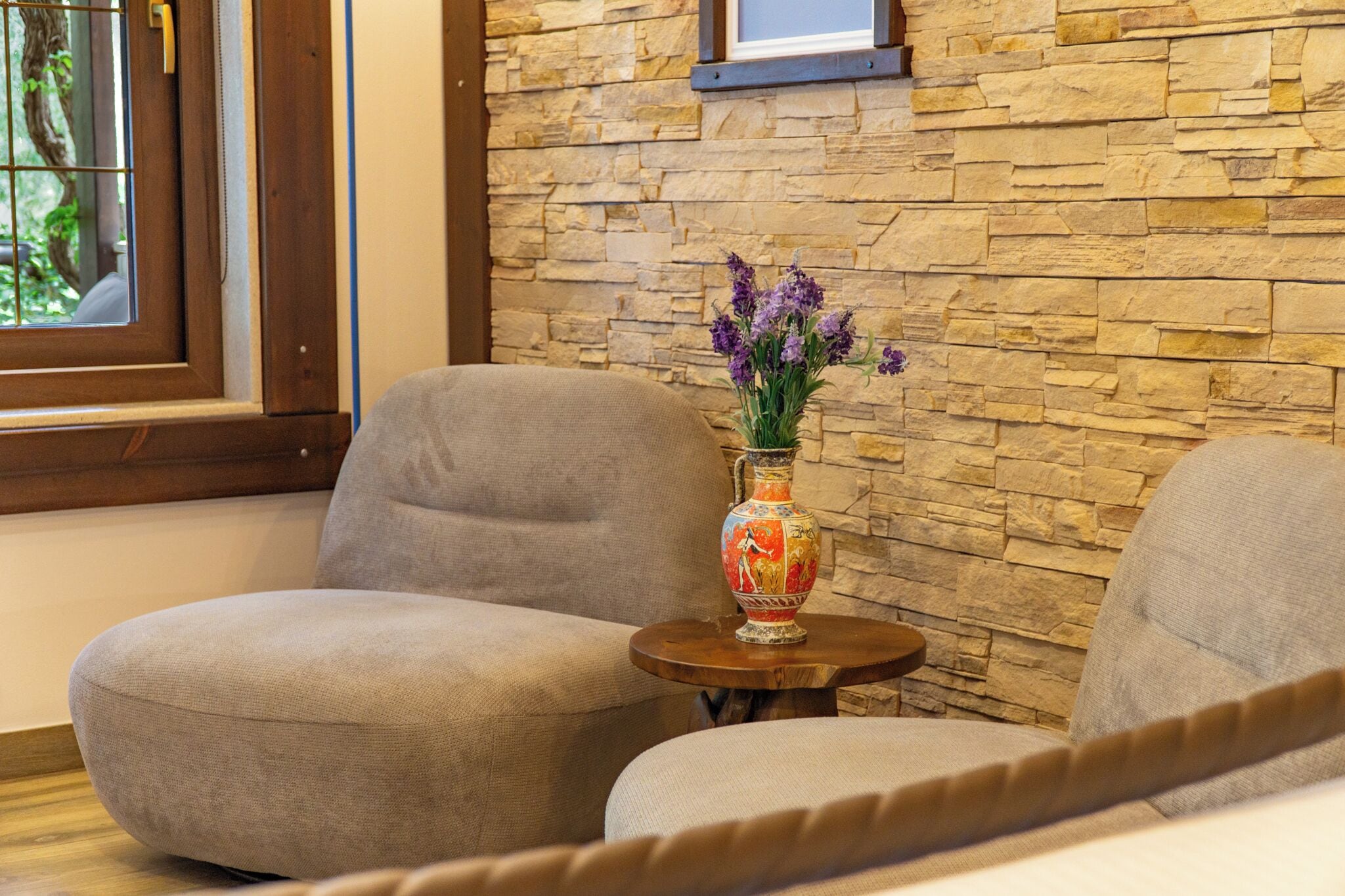 A room with a stone wall, two grey sofas, a small round wooden table, and a vase with flowers.