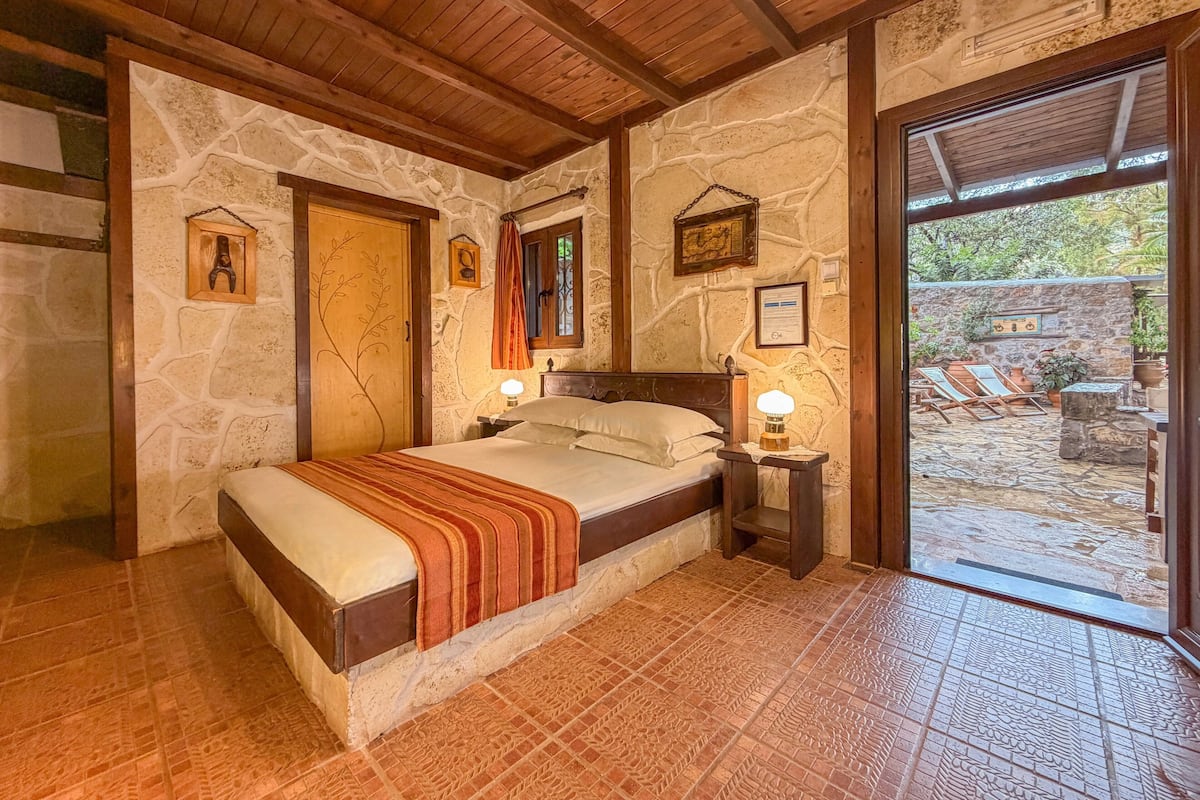 A bedroom with a stone wall, wooden ceiling, a bed with a blanket, bedside lamps, and a view of an outdoor area with lounge chairs.