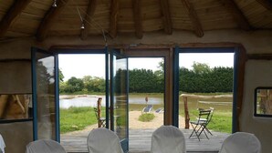 Una habitación con techo de madera y puertas de cristal amplias que dan a un espacio exterior con vistas a un lago y sillas cubiertas con telas blancas.