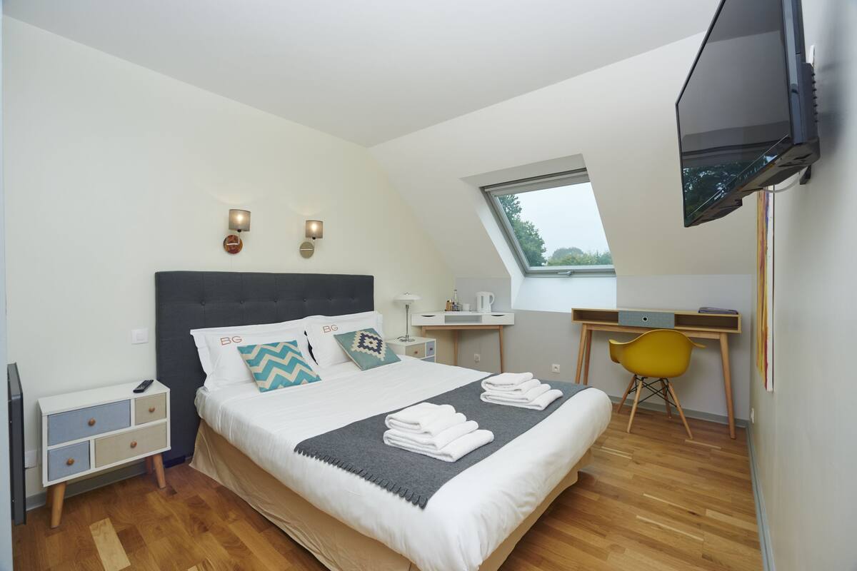 A modern bedroom with a bed, bedside table, TV, desk, chair, and a window.