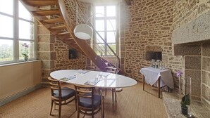 Un comedor con una mesa redonda, sillas, una escalera de madera, paredes de piedra y una chimenea.
