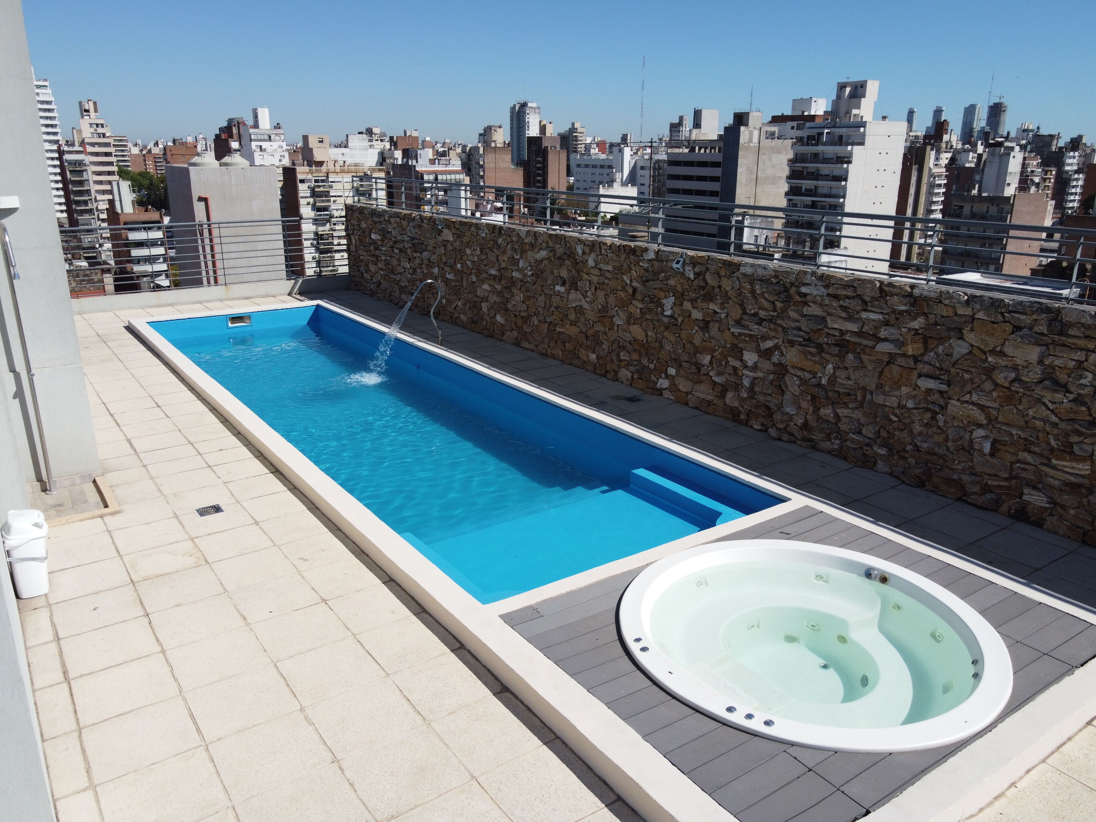 A rooftop swimming pool with a stone wall and a hot tub, surrounded by a cityscape.