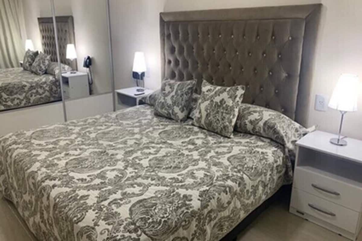 A bedroom with a large bed, a tufted headboard, patterned bedding, a nightstand with a lamp, and a mirror.