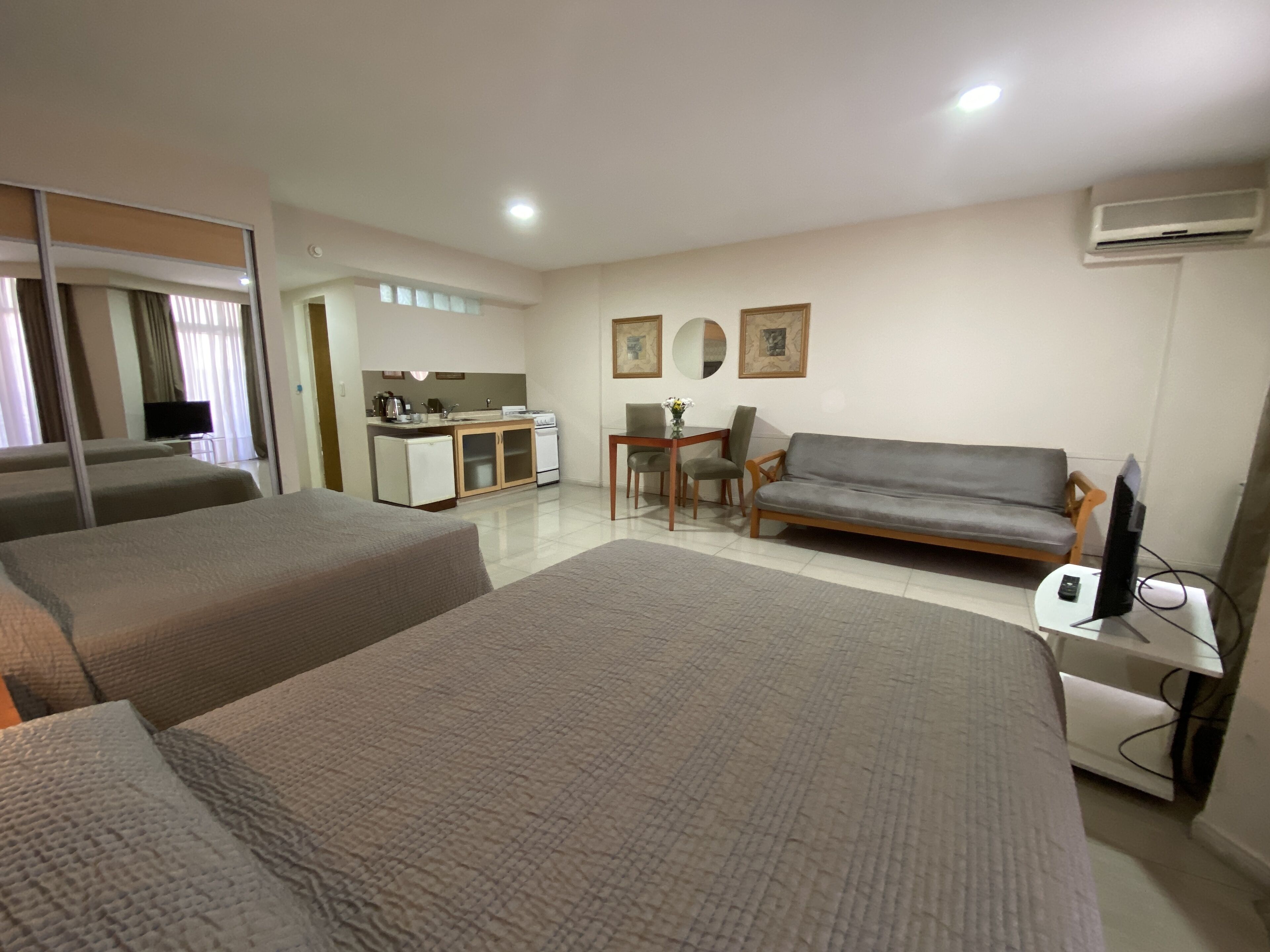 A hotel room with two beds, a sofa, a small dining table, a TV, and an air conditioning unit.