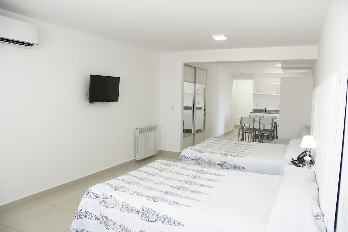 A hotel room with a bed, a TV, a radiator, and a view of the kitchen and dining area.