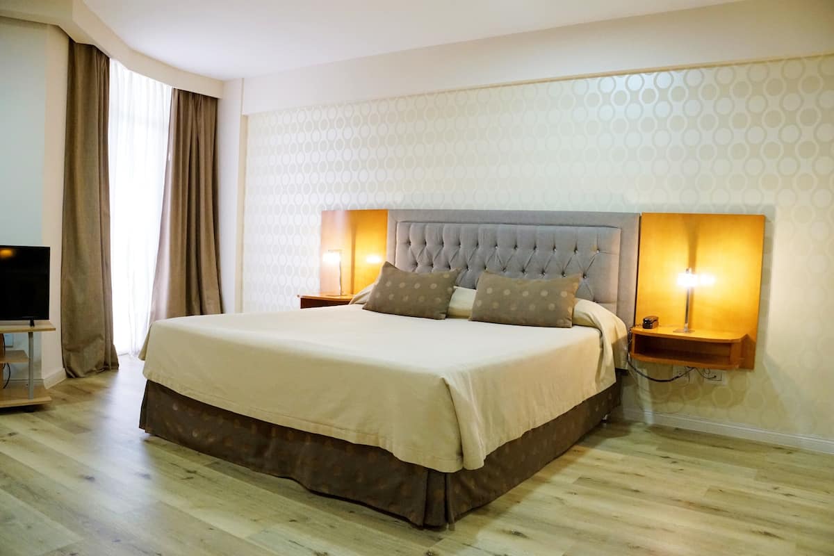 A neatly made bed with a grey headboard, two bedside lamps, and a TV on a stand to the left.