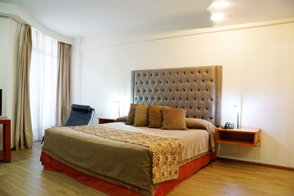 A neatly arranged hotel room with a large bed, a chair, a bedside table with a lamp, and a window with curtains.