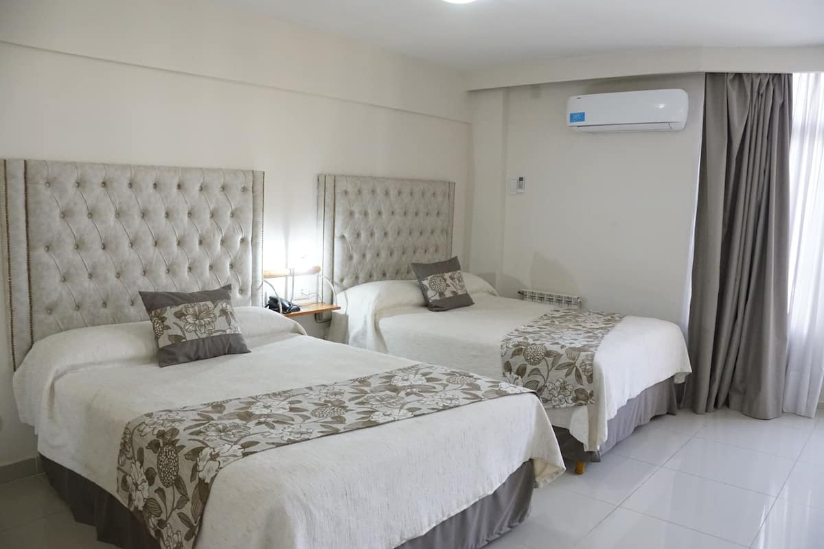A hotel room with two beds, each with a tufted headboard, a bedside table, and a large air conditioning unit on the wall.