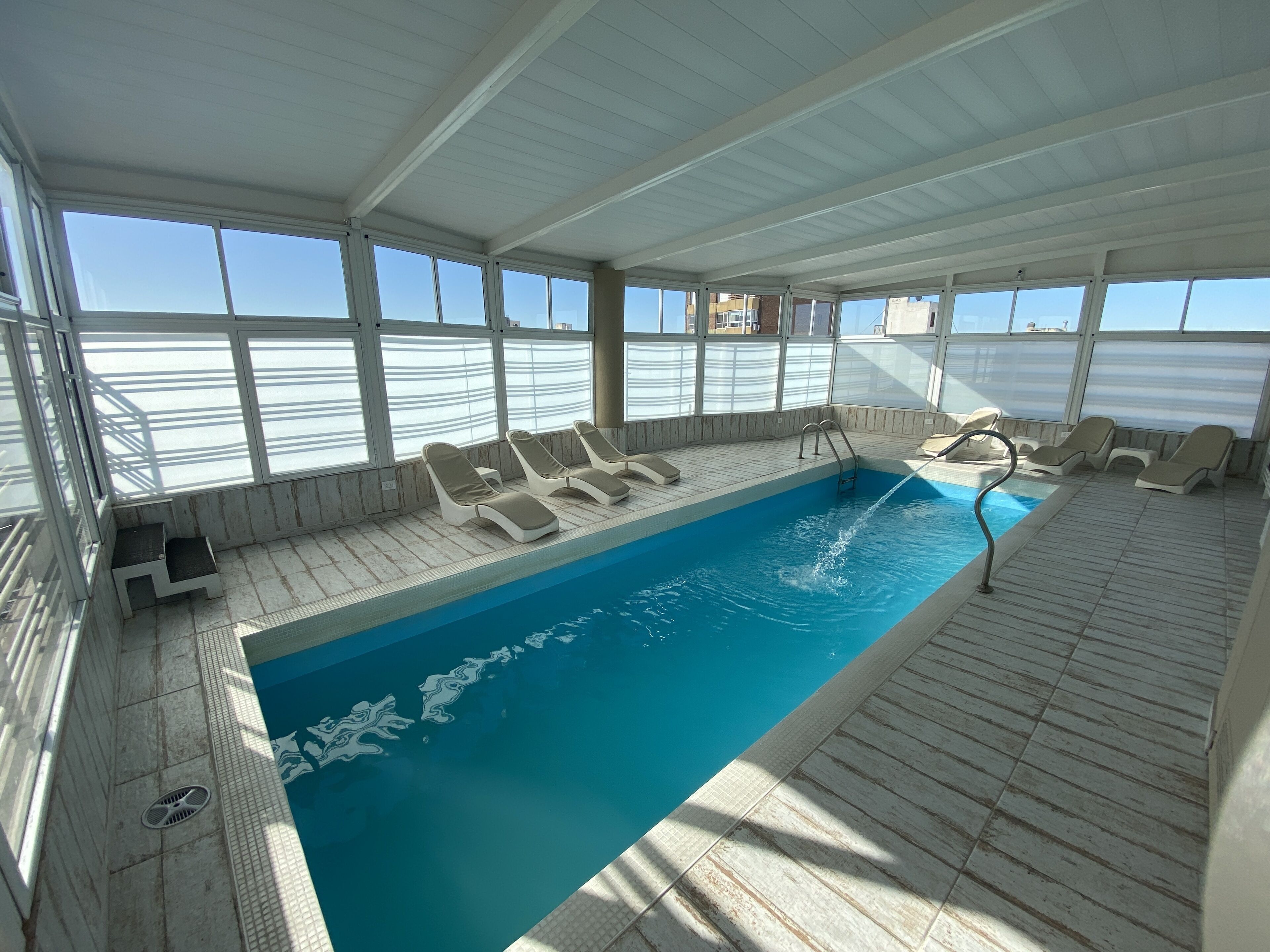 An indoor swimming pool with clear views of the sea through large windows, surrounded by lounge chairs and a wooden deck.