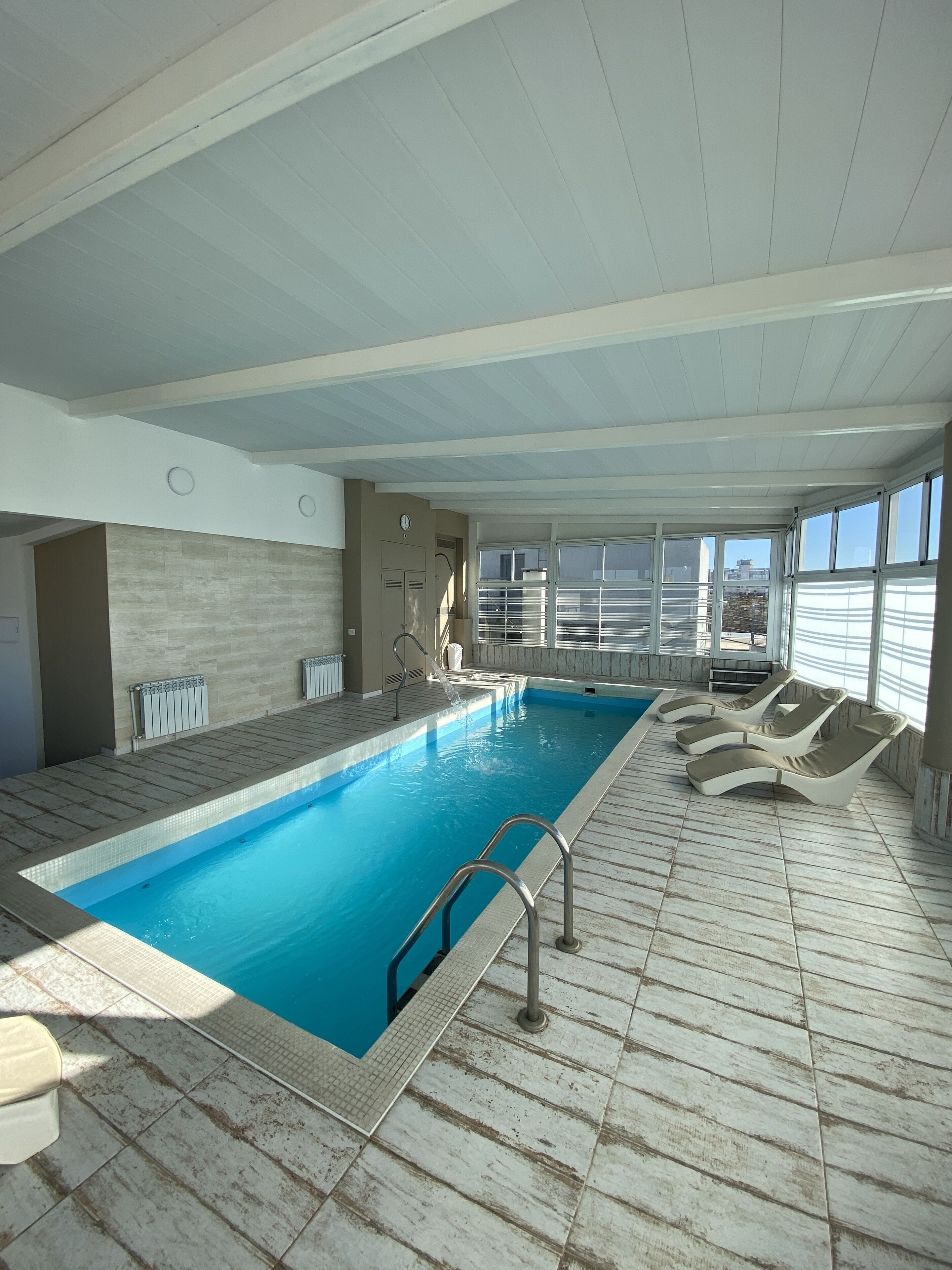 Una piscina interior con agua cristalina y azul, rodeada de sillones de descanso y un suelo de baldosas.