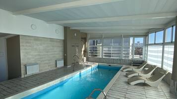 Una piscina interior con agua cristalina y azul, rodeada de sillones de descanso y un suelo de baldosas.