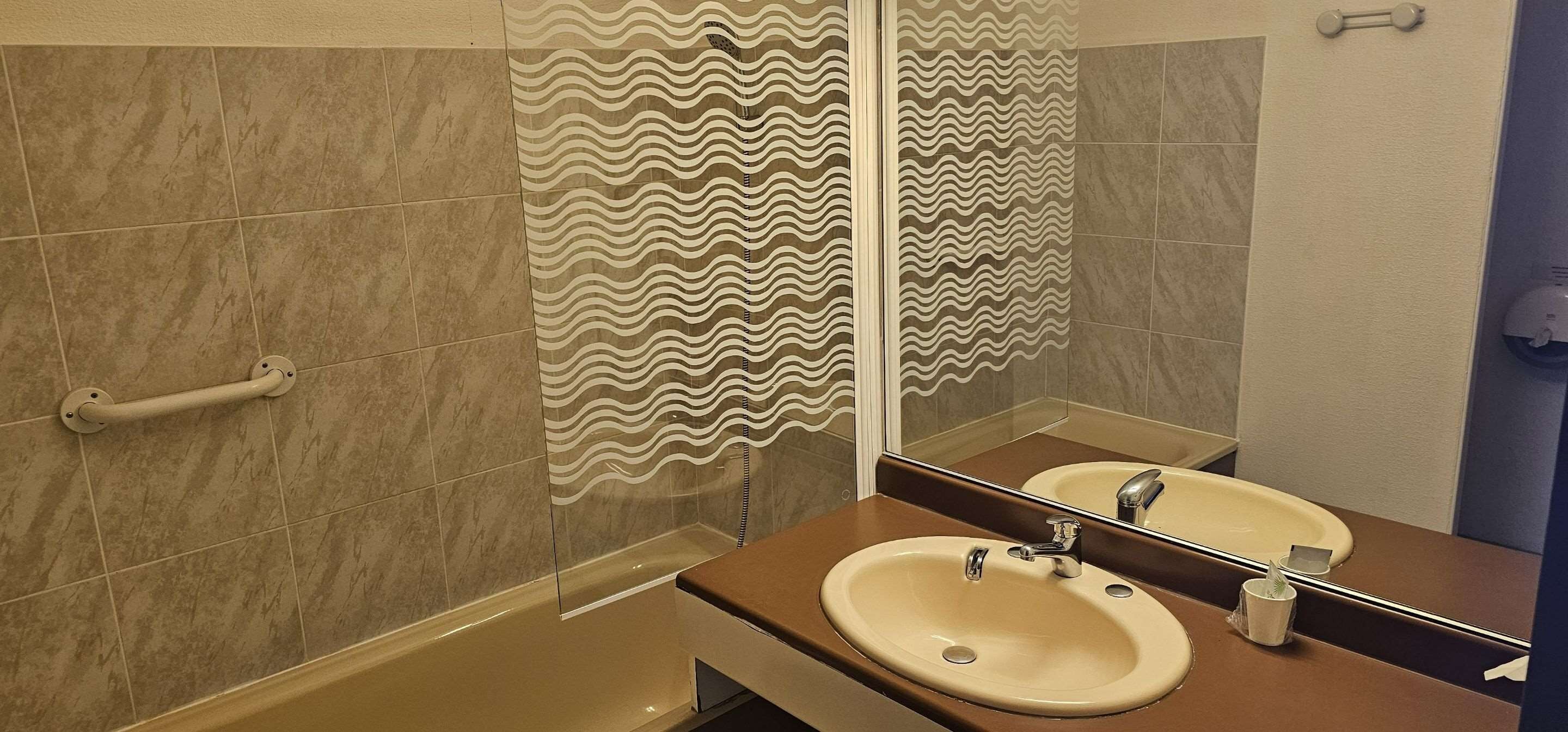 Un baño con un lavabo color crema, un espejo y una ducha con puerta de vidrio ondulada.
